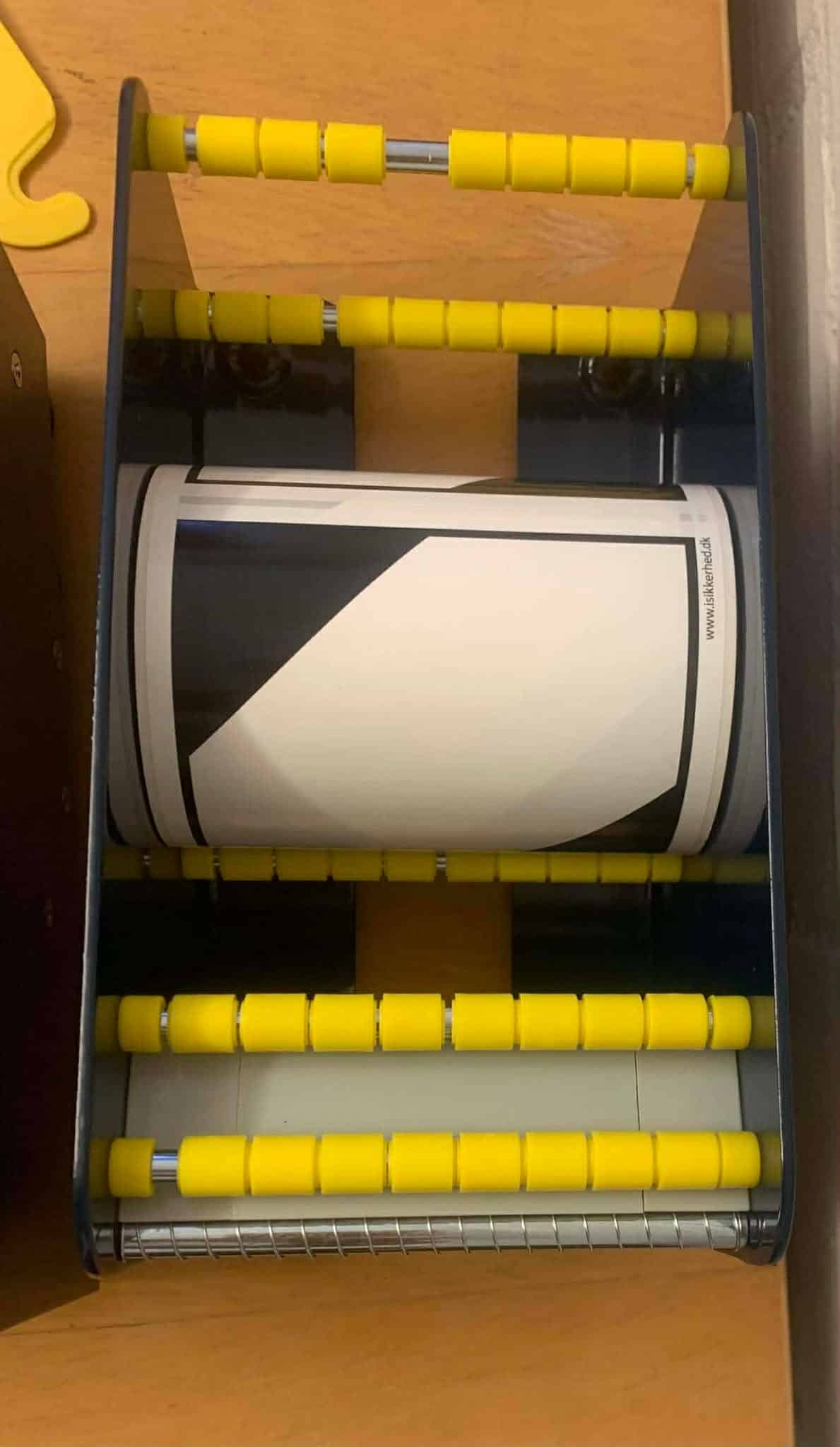 En etiketdispenser 120 mm med gule ruller holder en cylindrisk beholder på en træoverflade.