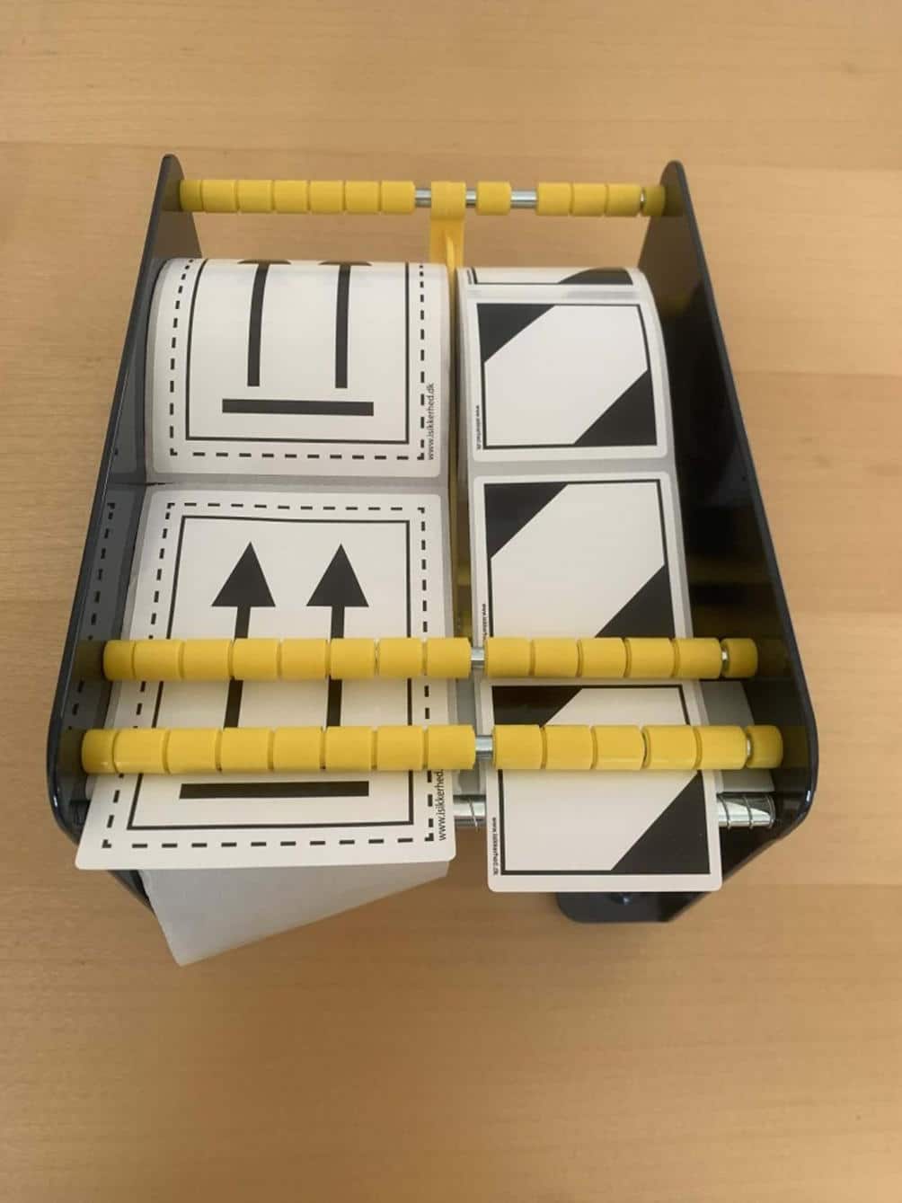 Etiketdispenseren 170 mm har plads til 4 ruller sorte pileetiketter og står på en lys træoverflade.