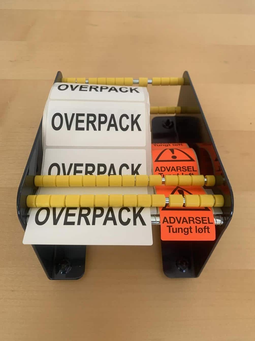En Label dispenser 170 mm holder OVERPACK-labels og orange ADVARSEL Tungt løft-klistermærker på en lys træoverflade.