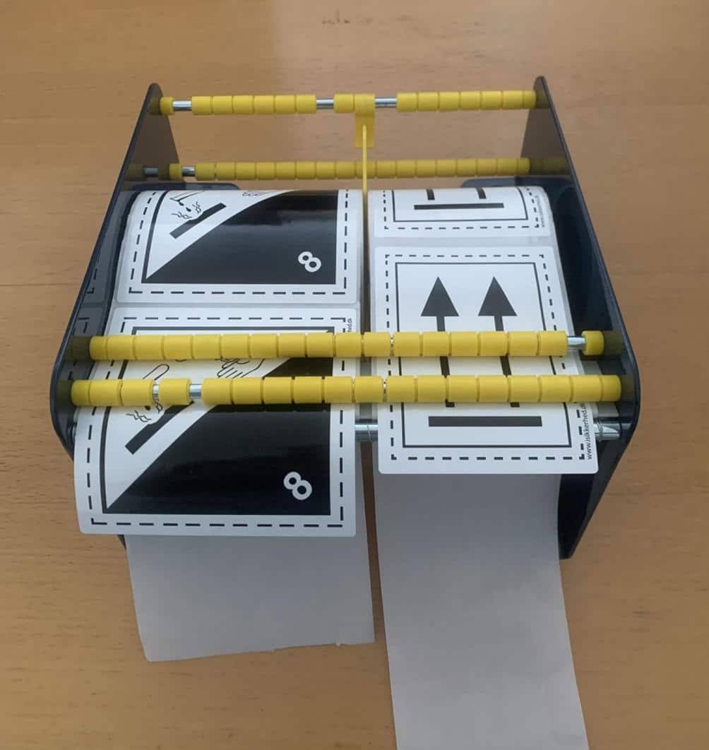 Etiketdispenseren 220 mm med gule ruller fremfører papirstrimler med kort og pile på en træoverflade.